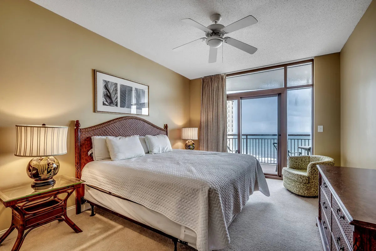 A serene master bedroom featuring a king-sized bed with plush bedding, large windows that frame an ocean view, decorative lamps beside the bed, and an accent chair positioned for relaxation. Soft, natural light fills the room, highlighting the tranquil color palette and inviting ambiance.