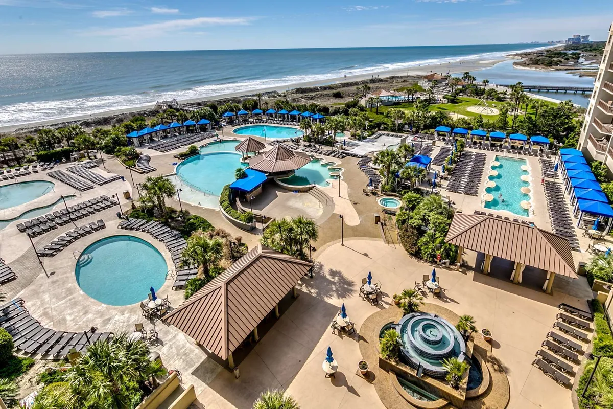 A stylish panorama of swimming pools with lounge chairs and shaded cabana structures set against an ocean view. The area is bustling with guests swimming, sunbathing, socializing, and unwinding, all under an inviting, sunlit sky.