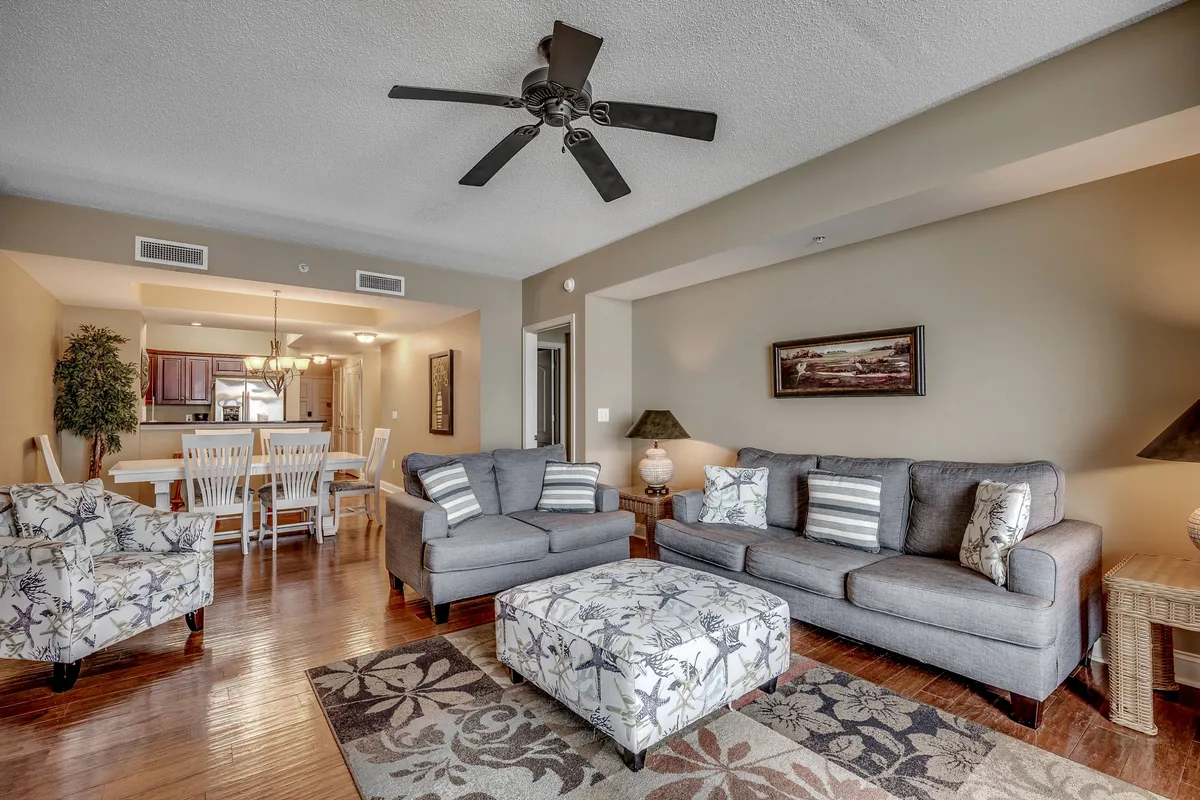 A wide view of an open-concept living and dining area, featuring a comfy sofa, decorative ottoman, elegant dining table, ceiling fan, and tasteful artwork. The layout encourages social gatherings and family relaxation, with subtle coastal touches in the décor.