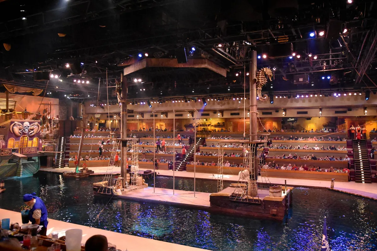 The photo shows a lively indoor theater with a ship-themed stage, decorative water features, and audience seating. Guests are enjoying a live performance, immersing themselves in a unique dining and entertainment experience.