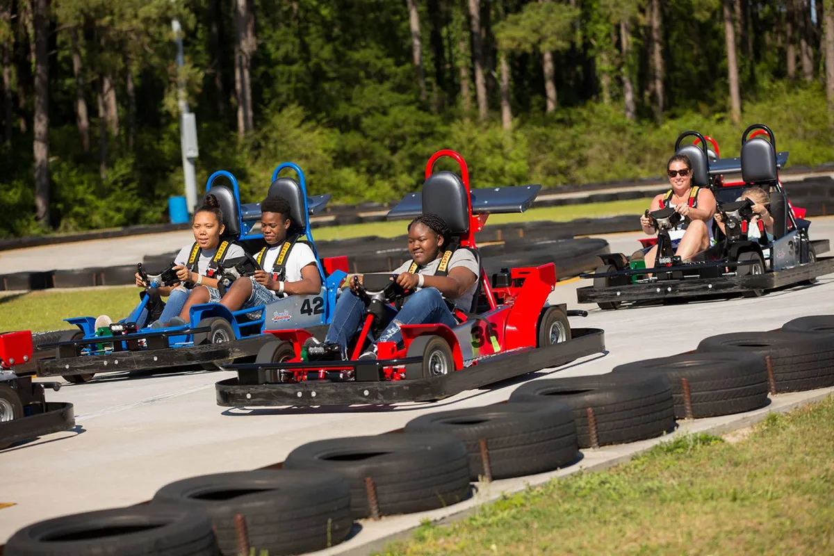 A lively go-kart track with families and friends racing around the bends, participants are seen in mid-action with expressions of excitement and fun. The track is bright and well-maintained, evoking the thrill and camaraderie of a classic family outing.