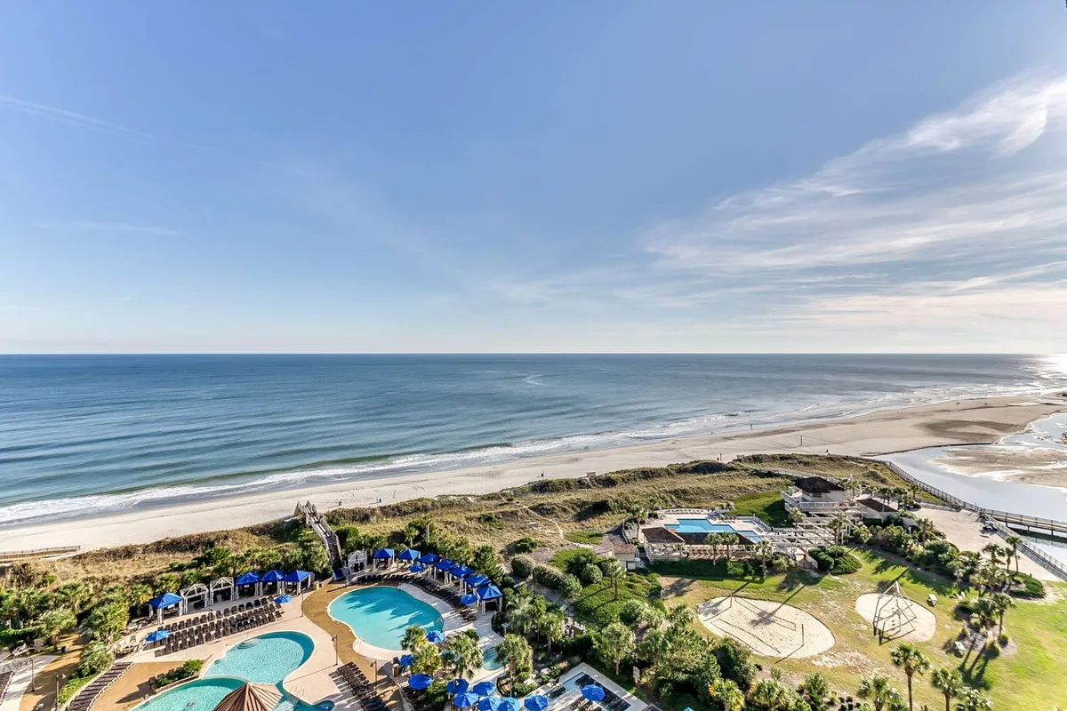A sweeping view over landscaped gardens reveals multiple swimming pools, a spacious beach area, and the glistening ocean beyond. The scene suggests a vibrant, sun-drenched day with guests enjoying swimming, sunbathing, and strolling between water features and the shoreline.