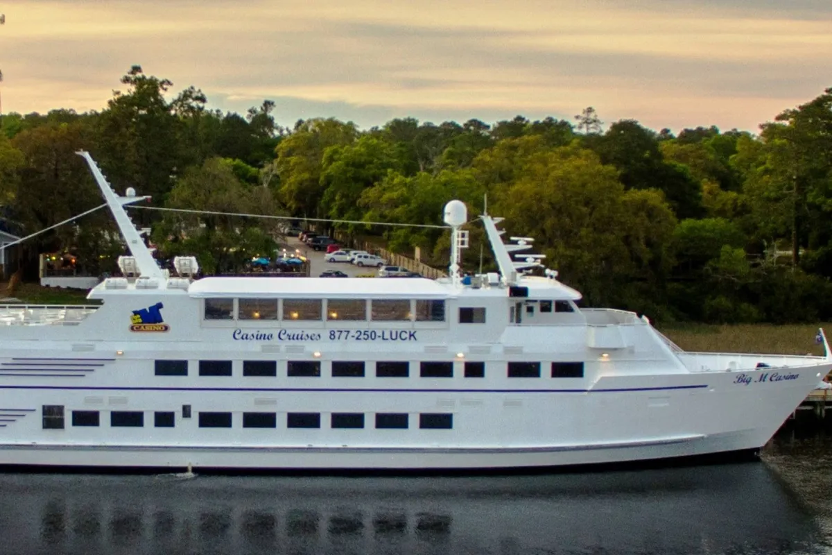 An exterior shot of a cruise ship docked amid lush greenery, its decks illuminated by the sunset reflected on the calm water. The scene hints at exciting entertainment and nightlife possibilities, with opportunities for casino gaming, dining, and enjoying scenic views under the stars.