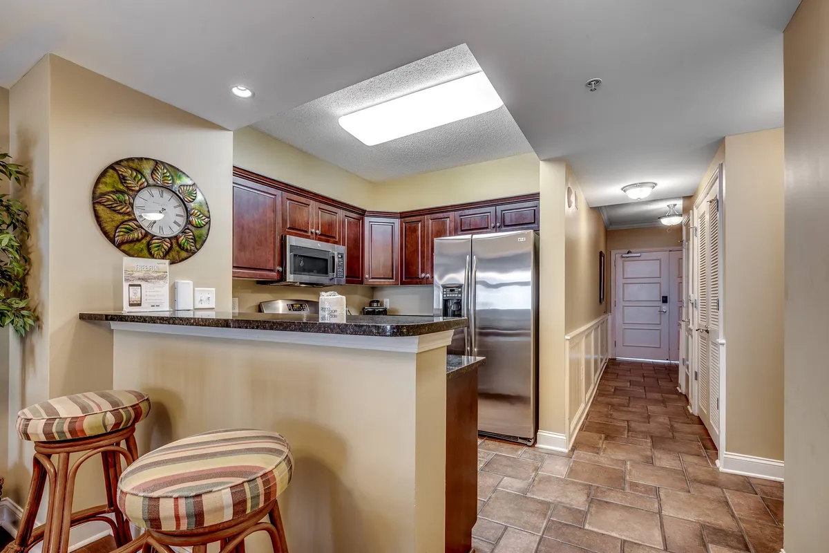 A cozy kitchen nook with a bar counter, kitchen cabinets, and stools, along with visible appliances and decorative touches. The setting invites socializing, whether it's a casual breakfast, a coffee chat, or prepping snacks, and suggests an easy transition from food prep to gathering around the counter.