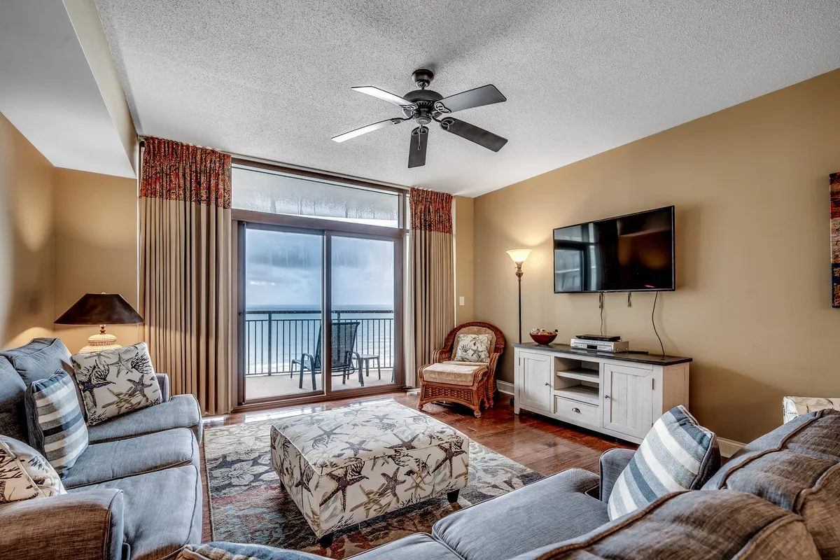This image captures a spacious and inviting living area with a comfortable seating arrangement, decorative ottoman, television unit, and large sliding glass doors that flood the room with natural light and offer glimpses of the outdoors. The décor is light and soothing, embodying a coastal-inspired ambiance that enhances the relaxing mood.