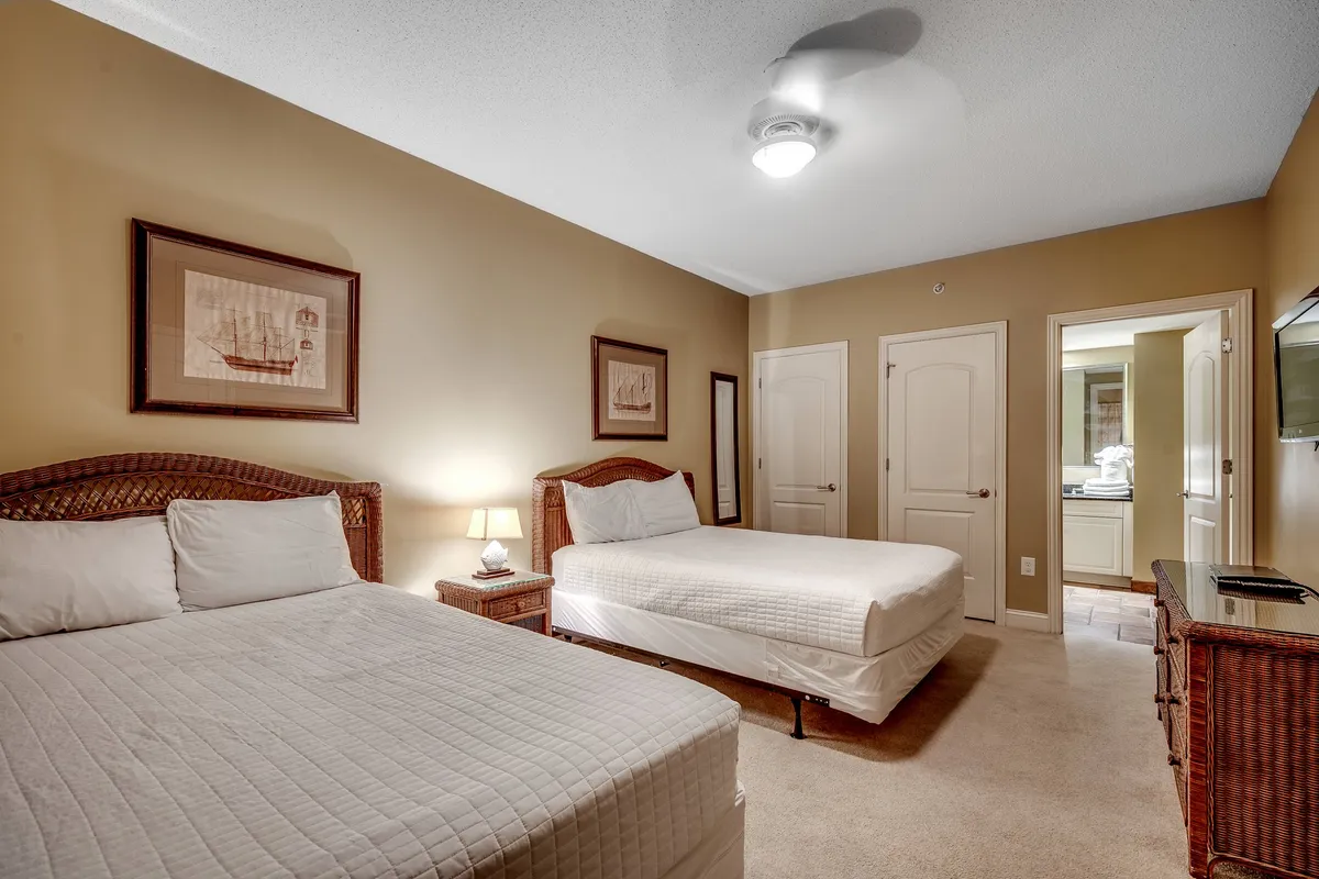 A comfortable second bedroom offering two neatly made beds with plush linens, a nightstand with a soft-glowing lamp, and framed artwork above the beds. The room exudes a quiet, restful ambiance, ideal for family members or guests.