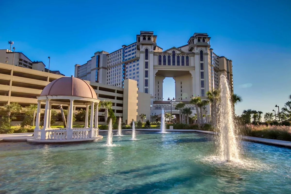 A beautifully landscaped exterior scene featuring a tranquil fountain, a charming gazebo, and elegant palm trees framing the resort entrance. The soft lighting evokes a serene, upscale atmosphere perfect for relaxation and romantic moments. The building's archway hints at the luxurious accommodations within, while the water elements add an inviting, peaceful touch.