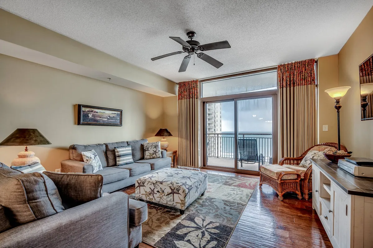The image features a stylish and inviting condo living room with a plush sofa, a chic coffee table, a cozy woven chair, and warm lighting from a lamp. The room opens up to a balcony with a view, inviting guests to relax indoors or step outside to enjoy sunsets. The setup hints at comfortable movie nights, lively gatherings, and serene moments overlooking North Myrtle Beach.