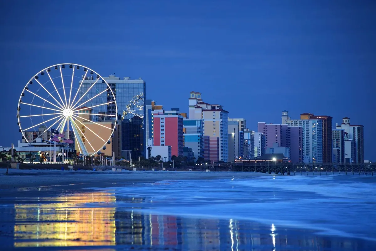 A vibrant beachfront scene with a Ferris wheel rising above colorful buildings, set against the backdrop of rolling ocean waves. The environment is festive and bustling, capturing the excitement and variety of attractions available nearby. Families are seen strolling and enjoying the lively beach atmosphere.