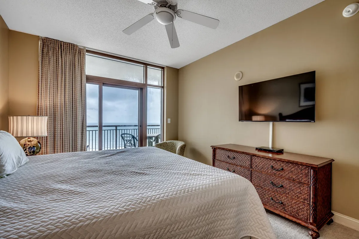 A cozy bedroom scene with a bed facing a window, a dresser topped with a television, and curtains softly framing the natural light. The space feels intimate and peaceful, inviting relaxation and quiet moments.