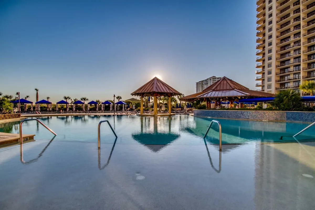 A sprawling outdoor pool area with sparkling blue water, surrounded by cabana structures and comfortable seating. The sun is setting over the pool, casting a warm glow and inviting atmosphere, perfect for family relaxation and fun. The scene suggests opportunities for swimming, sunbathing, and casual gatherings by the water.